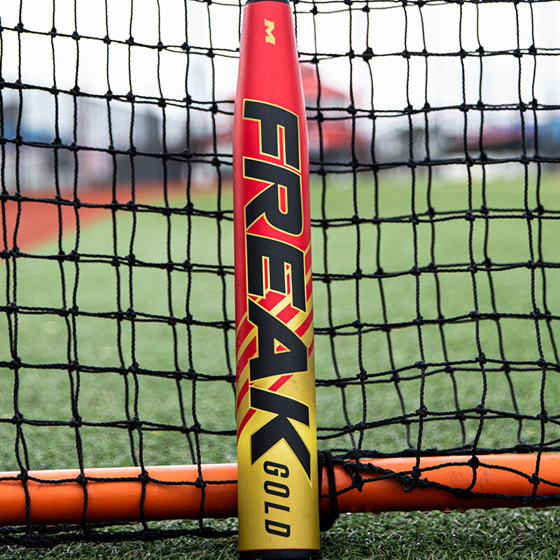 A red/gold Freak Gold USSSA bat leaning against the netting of a protective screen loading=