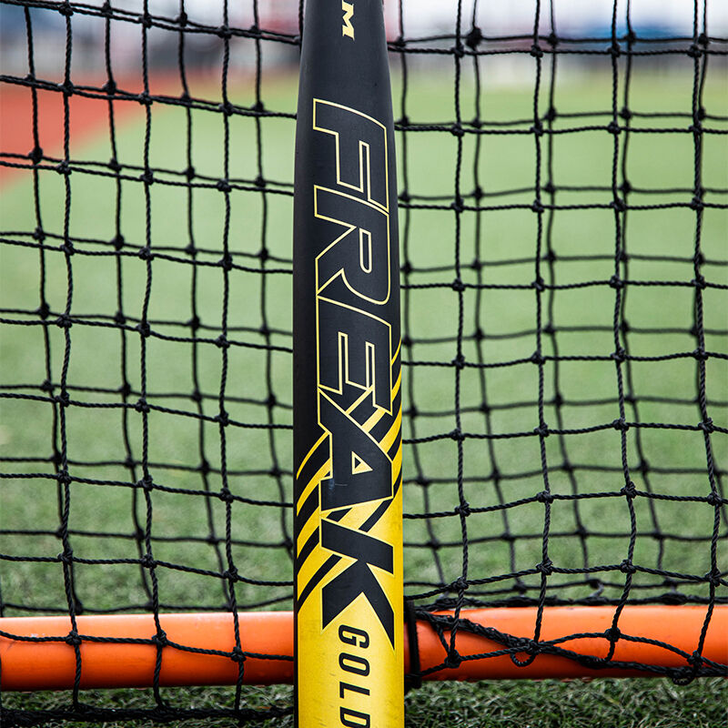 A black/gold Freak Gold Midload USA bat leaning against netting of a protective screen loading=